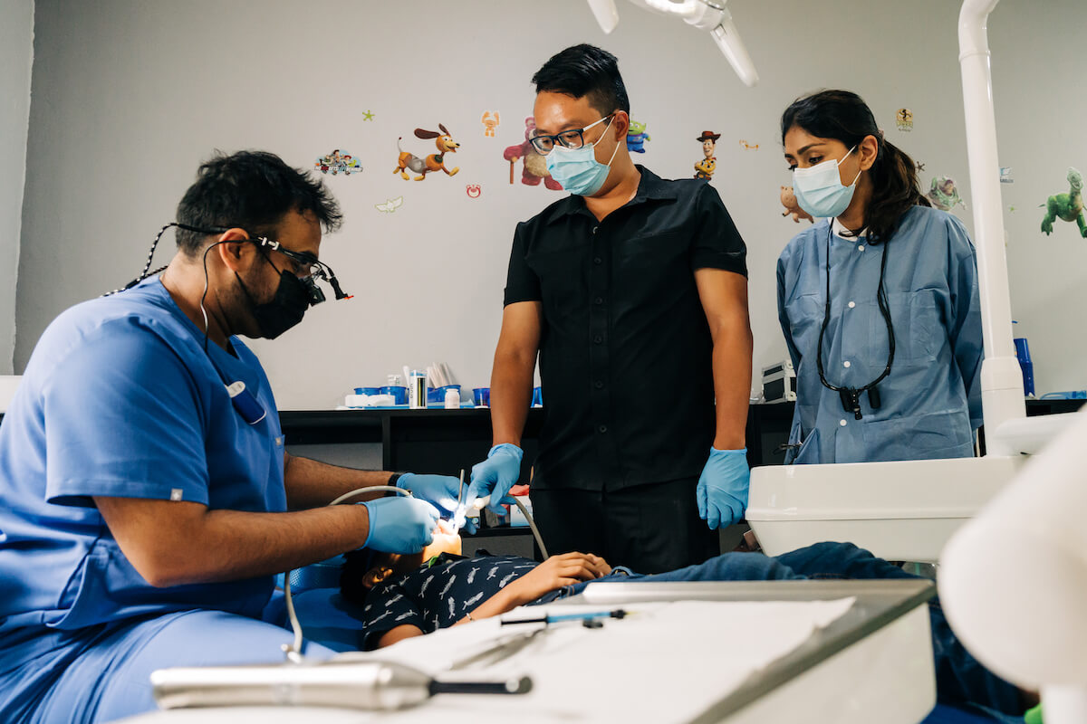 Two new dentists watch their mentor dentist perform a dental cleaning on a pediatric patient on a Humanitarian Trip. Learning on the job may be better for some new dentists vs AEGD vs GPR.