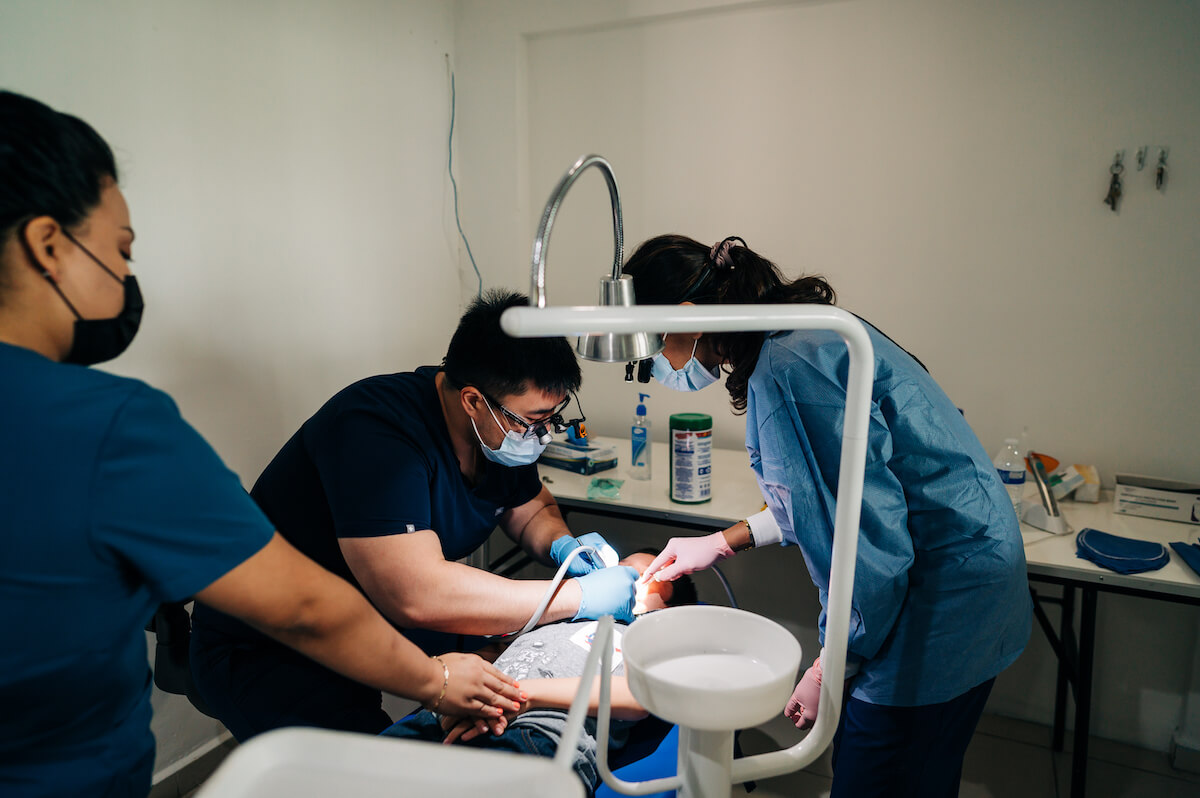 2 new dentists are overseen by a mentor dentist while treating a pediatric patient during a dental volunteer program.