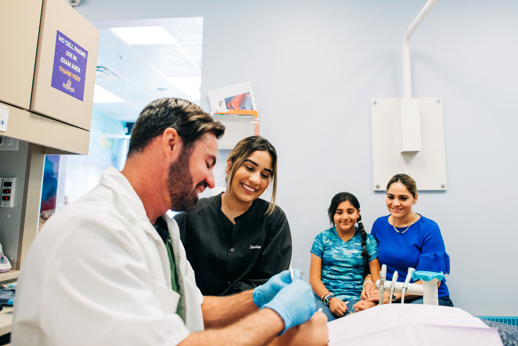 Pediatric dentist works on child as mom, sister, and assistant watch.